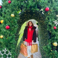 image of attractive, redheaded woman wearing black puffer jacket, aran jumper, red scarf and faux fur earmuffs, carrying brown paper shopping bags, walking on brick paved path under artificial conifer hedge arch, christmas ba