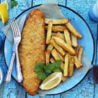 image of fish and chips (cod) garnished with parsley on turquoise plate with greaseproof parchment, knife and fork, lemon slice, bowls containing tartare sauce and mushy peas, bottle of vinegar, turquoise background, elevated