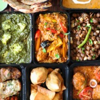 image of indian takeaway dishes in black plastic, single-use food containers, kadai chicken, chicken bhuna, palak paneer, samosas, murgh malai tikka, pink pickled onions, mint and coriander dip, lachha paratha, pindi chole a