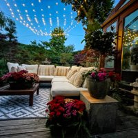 image of outdoor lounging area at night illuminated by string fairy lights, hardwood seating with cushions, wooden table top with flowering plant centrepiece, bonsai trees, japanese maples, landscaped oriental design garden, 