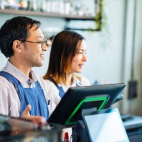 improve and increase the customer experience in coffee shops. shot of a male japanese coffee shop owner and a female waitress at a cashier counter and greeting customers as they enter the coffee shop. - junk food stock pictur