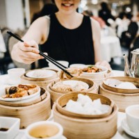 joyful asian woman picking up steamed beancurd roll with chopsticks and enjoying a variety of freshly made dim sum in restaurant - junk food stockfoto's en -beelden