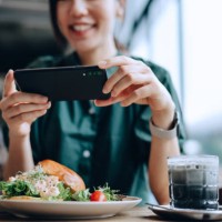 joyful young asian woman taking photos of delicious lobster salad brioche bun with bamboo charcoal black and white colour coffee with smartphone before eating it in cafe. eating out lifestyle. camera eats first culture - food