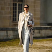Julia Haghjoo wears black sunglasses, silver large pearl earrings, a white latte silk knot neck blouse, a beige long wool coat, a white embroidered...