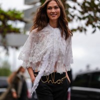 Karlie Kloss wears white sheer mesh lace flowy layered shirt, black high low-rise pants, gold Chloe belt, outside Chloe, during the Paris Fashion...
