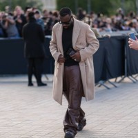 Maitre Gims wears black sunglasses, light brown blazer jacket, shiny dark brown leather pants, dark brown leather shoes, silver rings, outside Louis...
