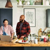 mature chinese woman and black mid adult man preparing food and laughing in kitchen preparing food with laptop on worktop - food stock pictures, royalty-free photos & images