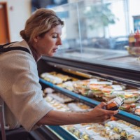 mature woman in casual clothes buying sushi at a supermarket, standing at a display of packaged food, focused on choosing, reflecting modern consumer lifestyle. - food stock pictures, royalty-free photos & images