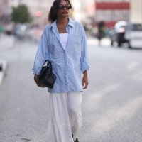 Maureen Adim is seen wearing black sunglasses with wide temples, hanging earrings with blue stone, an oversized blue and white striped shirt with...
