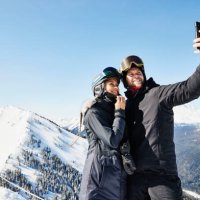 medium wide shot of smiling couple taking selfie while standing at the top of ski resort on winter morning - travel stock pictures, royalty-free photos & images