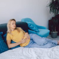 middle-aged woman holds her hands on her stomach while lying on a bed at home. pain, negative emotions and suffering. - junk food stock pictures, royalty-free photos & images