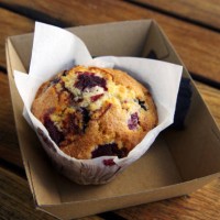 mixed berry, white chocolate and coconut muffin in waxed paper in a cardboard tray on a wooden outdoor table - junk food stock pictures, royalty-free photos & images