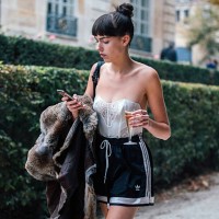 Model Kye Howell checks her phone, drinks champagne, and wears a white corset top and black sporty Adidas shorts after the Dior show at Musee Rodin...