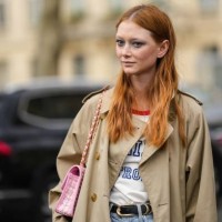 Model wears a beige long belted trench coat, blue denim pants, a pink and white checkered print pattern tweed shoulder bag from Chanel, a black shiny...
