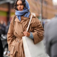 Model wears a blue knit scarf over the head as a hood / balaclava, a brown trench coat, holds a paper shopping bag outside Ulla Johnson , during New...