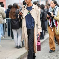 Model wears a dark blue pullover, a beige trench coat, black pants, brown and black leather shoes, a shopping bag, leather shoes, outside Nina Ricci,...
