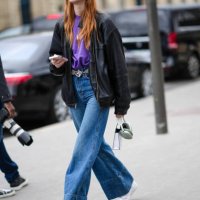 Model wears a purple t-shirt , a black leather jacket , a belt, blue flared denim pants / jeans , white sneakers shoes , outside Chanel, during the...