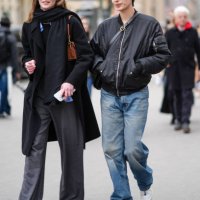 Model wears a scarf, a black coat, gray flared pants ; a model wears a black bomber jacket, blue denim pants / jeans , white sneakers shoes, outside...