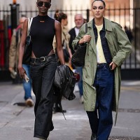 Model wears a sleeveless black top with a high neckline, paired with high-waisted dark denim jeans. Accessories include large black sunglasses and a...
