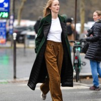 Model wears a white t-shirt, a green long coat with black quilted lapels, brown flared corduroy pants, sneakers shoes, outside Chanel, during the...