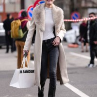 Model wears black eye shadow, white long sleeve shirt, cream wool coat with light gray faux fur collar, a black denim jean skinny pants, shiny black...