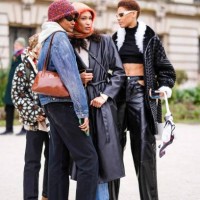 Model wears black sunglasses, a white hoodie sweater, a blue faded denim jacket, a camel shiny varnished leather shoulder bag, black denim pants,...