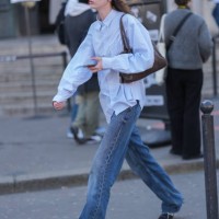 Model wears black sunglasses, light blue buttoned up long sleeve shirt, shiny dark brown leather bag, navy blue washed loose denim jean pants, black...