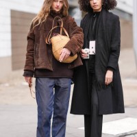 Model wears dark brown sunglasses, dark brown suede jacket, dark brown sweater, navy blue relaxed trouser pants, light brown woven basket, shiny...