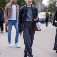 Model wears dark gray sweater, white t-shirt, shiny black oversized leather jacket, black leather belt, dark gray loose pants, shiny black loafer...
