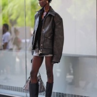 Model wears gold earrings, gold necklace, black shirt, dark brown oversized blazer jacket in leather, light gray mini skirt, shiny black pointed toe...