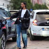 Model wears white cotton t-shirt, black oversized baggy leather jacket, shiny black woven leather bag, black leather belt, light blue denim jean...