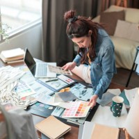 new work consulting on smartphone asian adult female woman fashion designer cloth tailor working for new collection sketch drawing choosing new color swatch fabric sample at her desk in her design studio home studio - fashion