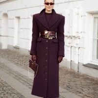 Nina Sandbech wears a burgundy top, long burgundy coat with multiple belts, burgundy bag, burgundy leather gloves and pointy burgundy shoes outside...