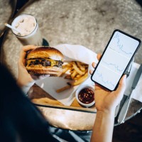 over the shoulder view of busy young woman checking financial trading data on smartphone while having her meal, beef and cheese burger with fries in a restaurant - junk food stock pictures, royalty-free photos & images