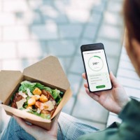 over the shoulder view of young asian businesswoman using fitness plan mobile app on smartphone to tailor make her daily diet meal plan, checking the nutrition facts and calories intake of the food while having meal outdoors 