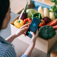 over the shoulder view of young asian woman doing home delivery grocery shopping online with mobile app device on smartphone at home, with a box of colourful and fresh organic vegetables and fruits on the table - food stock p
