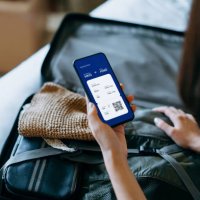 over the shoulder view of young asian woman using smartphone, checking digital flight ticket and boarding pass on device screen while packing a suitcase on bed for a trip. planning for travel. travel and vacation concept - tr