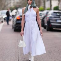 Penelope Guerrero wears black sunglasses, dark green pattern/print head scarf, white halter pleated maxi dress, cream leather bag, white heeled boots...