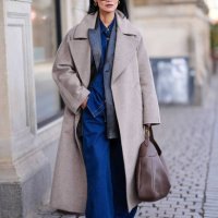 Pornwika Spiecker wears sunglasses , earrings, a gray long wool / trench coat, a black leather jacket , blue flared denim jeans pants, a large...
