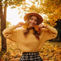 portrait of a beautiful young woman in a hat and a yellow sweater having fun and holding an autumn leaf enjoying solitude walking through the foliage in an autumn park outdoors in autumn. a pretty smiling girl in fashionable 