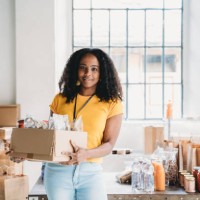 portrait of a mixed race volunteer woman holding a cardboard box of food and drink at the food bank - food stock pictures, royalty-free photos & images