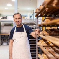 portrait of a senior baker in apron standing by bread rack - food stock pictures, royalty-free photos & images
