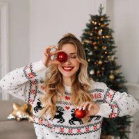 portrait of a young cheerful woman with red lips and curly hair in a knitted sweater laughing and holding a red christmas ball covering her eyes with it against the background of a christmas decorated christmas tree at a holi