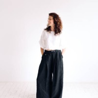 portrait of laughing beautiful authentic young woman with curly brown hair and brown eyes standing against white background with hands in pants pockets. she is wearing loose linen casual clothing. concept of natural beauty - 