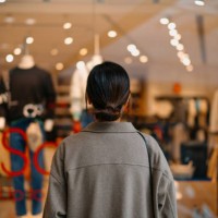 rear view of young asian woman standing outside a boutique looking at clothings through shop window while shopping in shopping mall. window shopping. sale season. seasonal shopping and consumerism concept - fashion stock pict