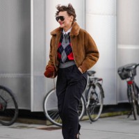 Renia Jaz wears aviator sunglasses, earrings, a brown aviator jacket with fluffy inner lining , an argyle pattern colored pullover , suit flared...