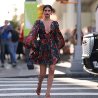 Sara Sampaio is seen wearing golden earrings; a mini dress with puffy long sleeves, a deep neckline and a blue/red/green floral print; a black...