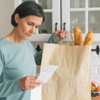 senior adult female worried while looking at receipt from grocery store. rise in price of products concept. buying consumer goods during inflation. facing economic downturn. kitchen unit and paper bag on background, horizonta