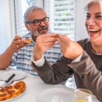 senior couple laughing while eating pizza at home - junk food stock pictures, royalty-free photos & images