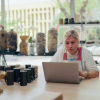 senior female potter working on laptop in her pottery shop - home decoration stock-fotos und bilder